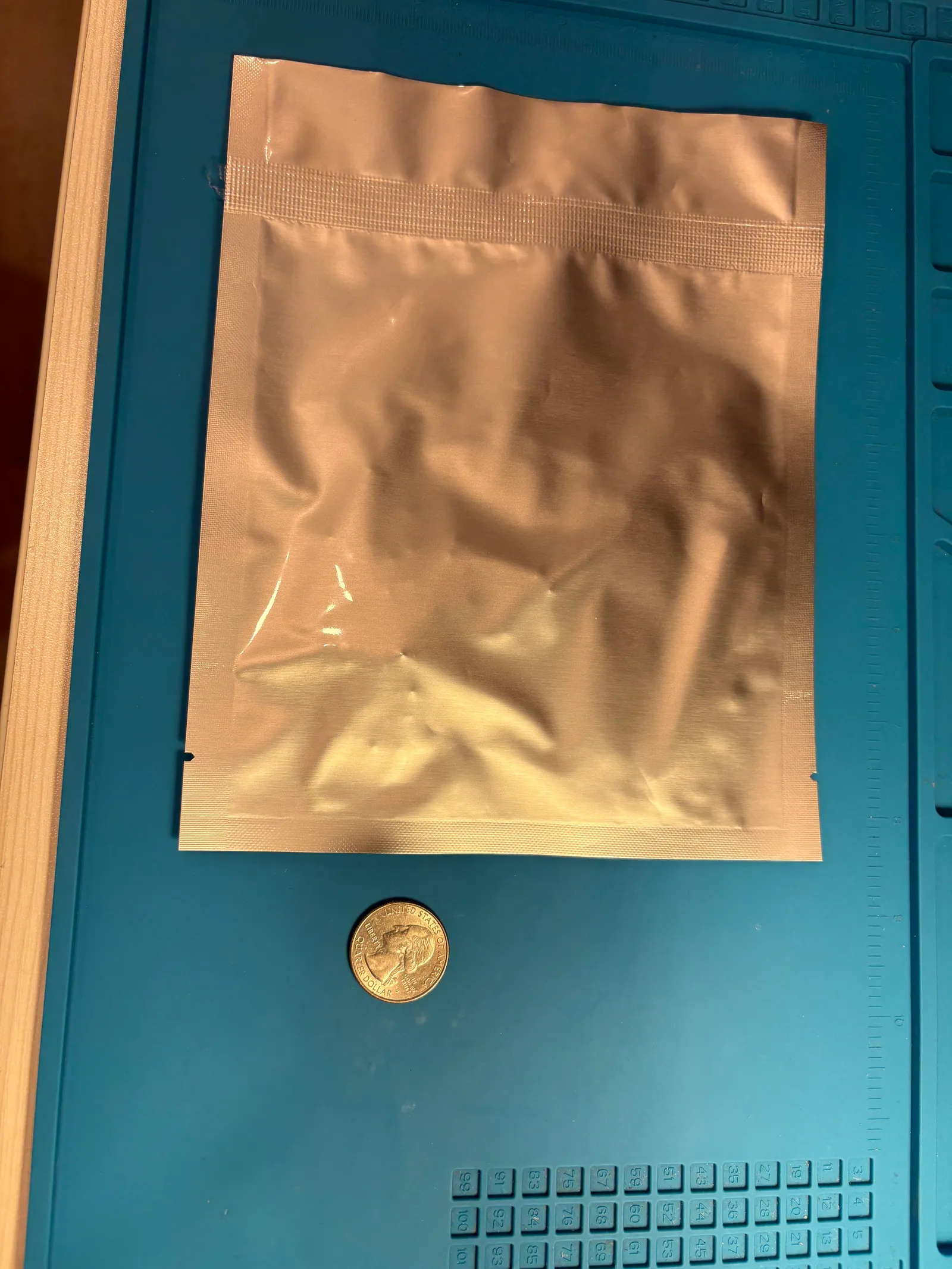 A sealed silver moisture-barrier bag on a desk mat, with a quarter beside it for scale.