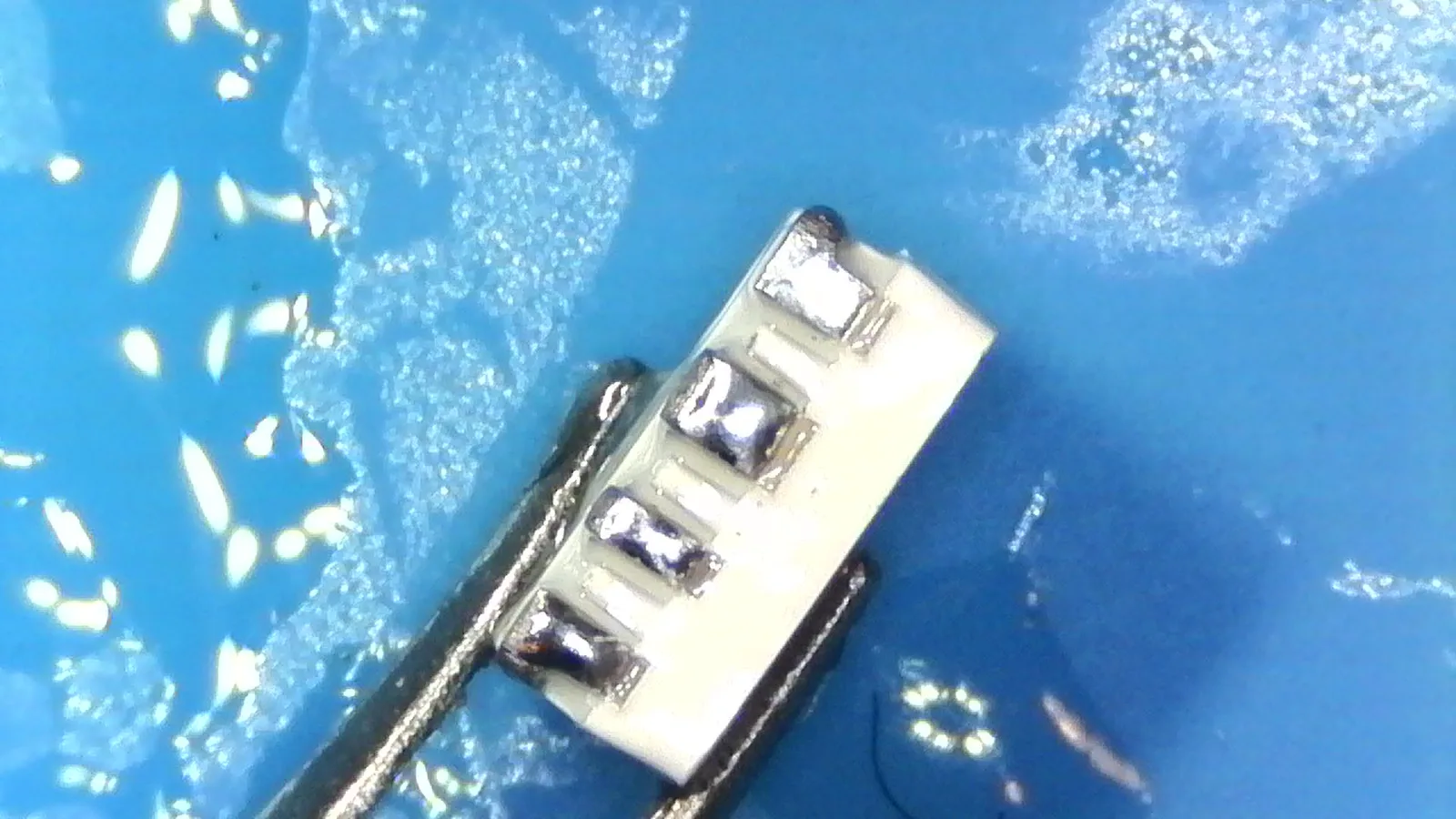 Microscope view of an LED held in tweezers, each of its four pads tinned with a bead of solder.